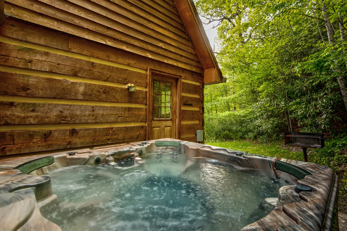 An inviting hot tub set just outside the log cabin, surrounded by lush greenery. The scene reflects a tranquil setting for soaking under the stars or relaxing after a day of activities.