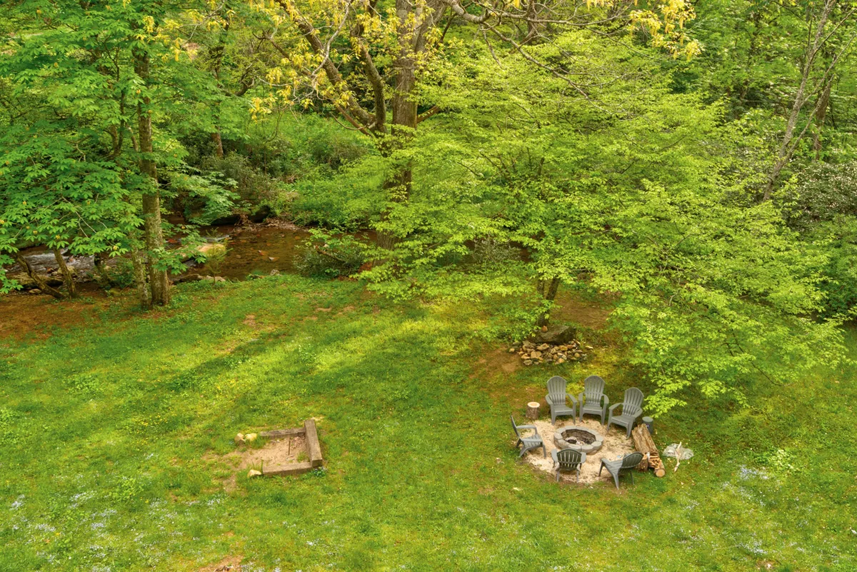 A cozy fire pit area with Adirondack chairs sits beside a gently flowing stream, surrounded by tall trees and greenery. The inviting seating arrangement encourages gatherings, storytelling, or stargazing, with the creek providing a soothing backdrop.