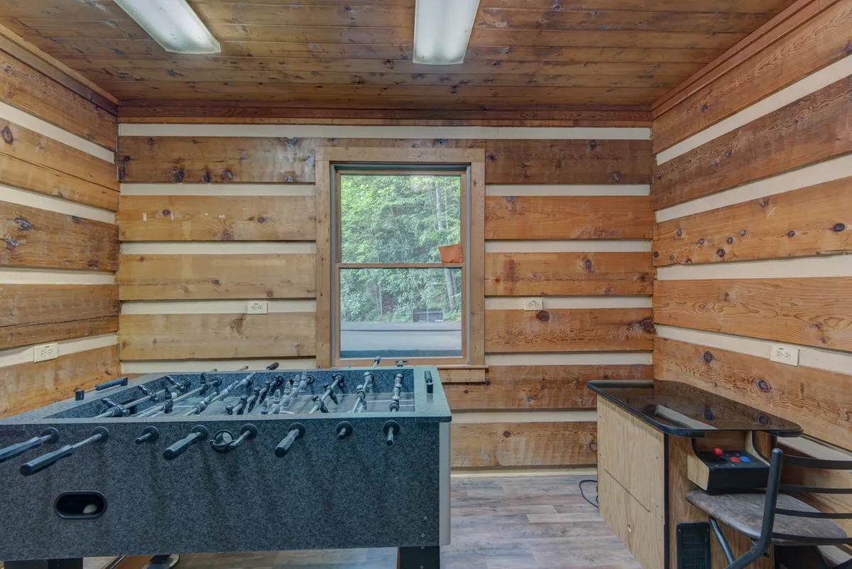 A lively interior scene centered on a foosball table, set against warm wooden walls with a window framing outdoor views. The room feels fun and welcoming, ideal for indoor entertainment on rainy days or evenings.