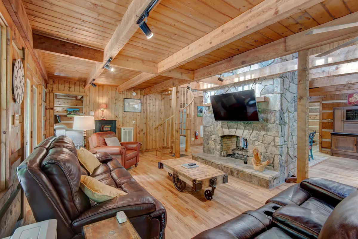 A plush, inviting seating area is arranged around a tall stone fireplace, with leather sofas and a coffee table making it ideal for movie nights or storytelling sessions. The wooden beams and warm lighting add to the room’s coziness, while a TV mounted above the fireplace hints at entertainment options.
