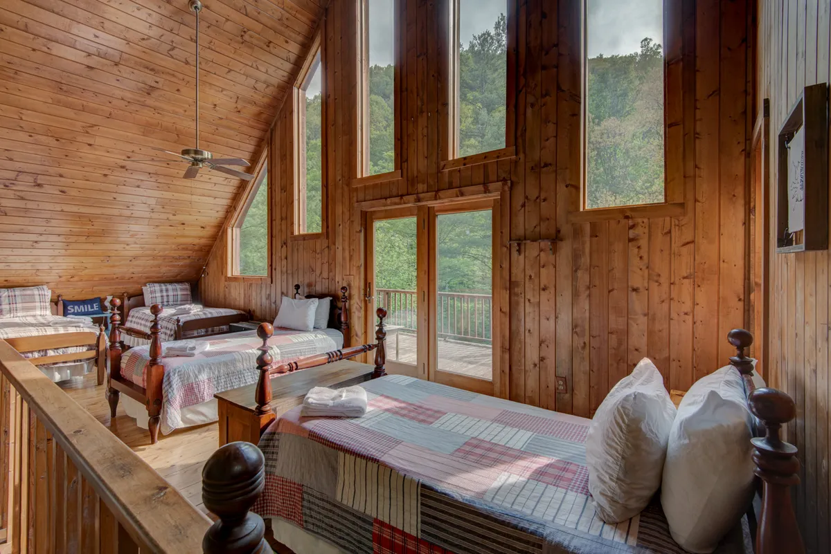 A bright, cozy sleeping area with large windows, wooden beams, and bunk beds with plaid bedding. The arrangement is clearly communal, evoking the feel of a camp-style sleepover for kids and families, with plenty of natural light.