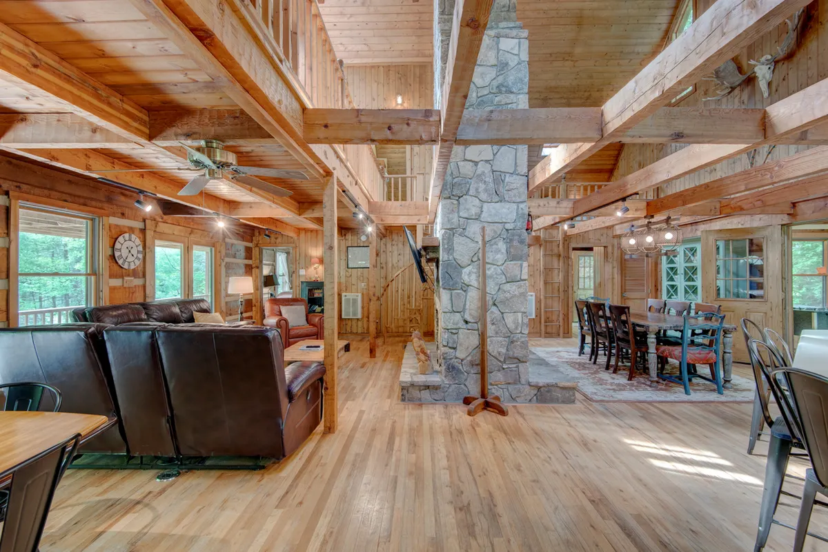 This image features a warm, welcoming great room centered around a striking stone fireplace. The space is framed by wooden beams and flooded with natural light from large windows. In the foreground, a spacious dining table is set for a gathering, and inviting seating is arranged near the fireplace, creating a perfect environment for cozy evenings and social activities.