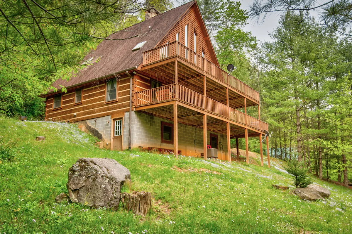 A wide view of the cabin’s wooden balcony, log construction, and surrounding trees sets a welcoming mountain retreat scene. The balcony looks ideal for group gatherings, outdoor dinners, and soaking up sunset or starlit views.