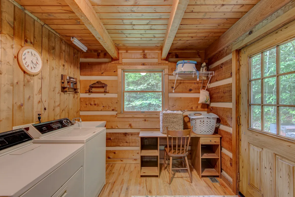 A practical and inviting laundry area with washing machines, a wooden chair, and a window overlooking greenery. Sunlight enhances the space, making daily tasks feel more pleasant and convenient.