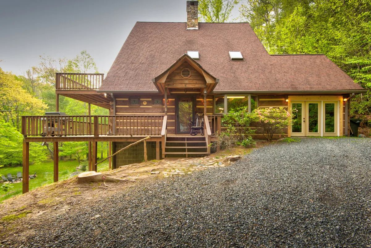 The hero image showcases a classic log cabin nestled among trees, with inviting front steps leading up to a spacious deck. The setting exudes tranquility, framed by natural greenery and the soft light of the Blue Ridge Mountains. The deck is perfect for lounging, sipping coffee, or stargazing, setting the scene for relaxation and connection with nature.