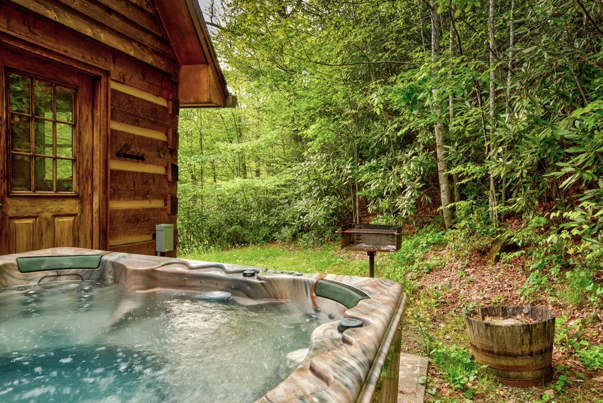 A rustic log cabin nestles among abundant greenery, featuring a private hot tub and a grill on the porch area. The hot tub looks out toward the woods, creating a perfect spot to unwind surrounded by nature. The scene suggests opportunities for soaking after outdoor activities and highlights amenities for both relaxation and outdoor dining.