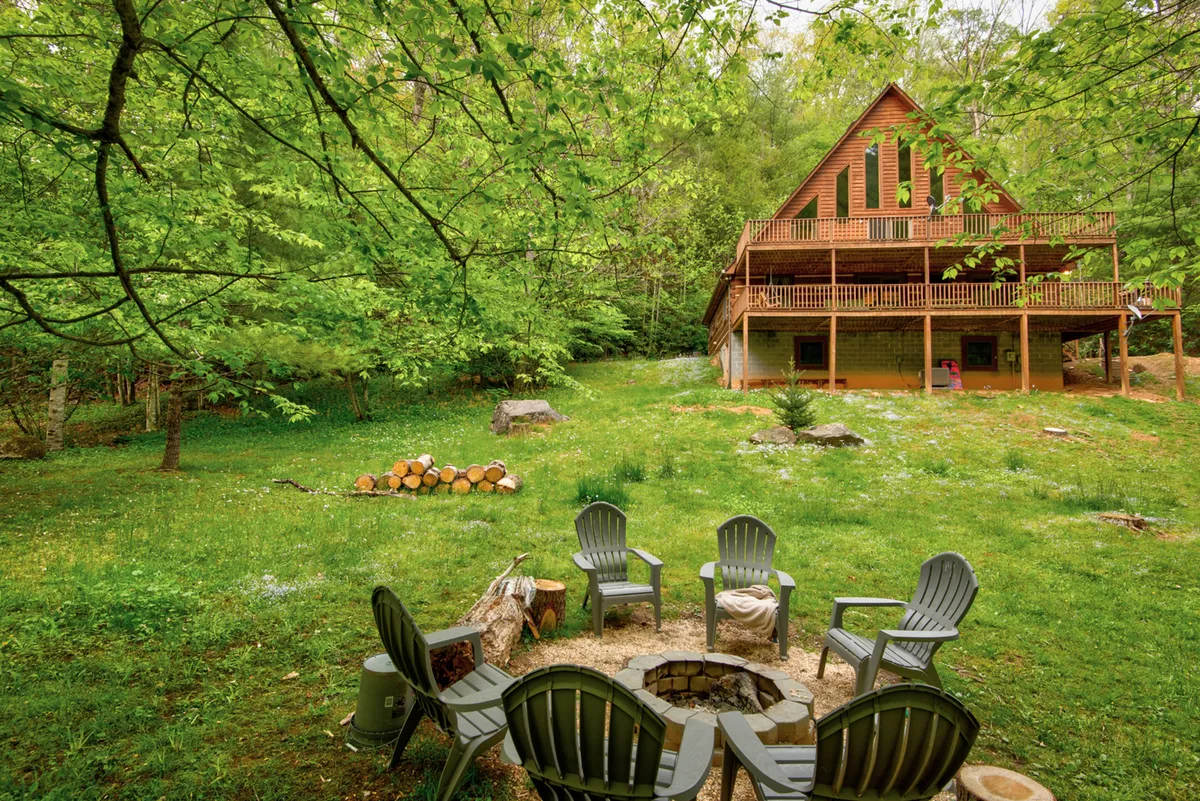 A cozy exterior scene showing a wooden cabin surrounded by lush greenery and featuring a fire pit circled by Adirondack chairs, inviting guests to gather outdoors. The setting conveys a welcoming, communal atmosphere with clear views of nature all around, perfect for pets and their owners to relax together after a day of adventure.