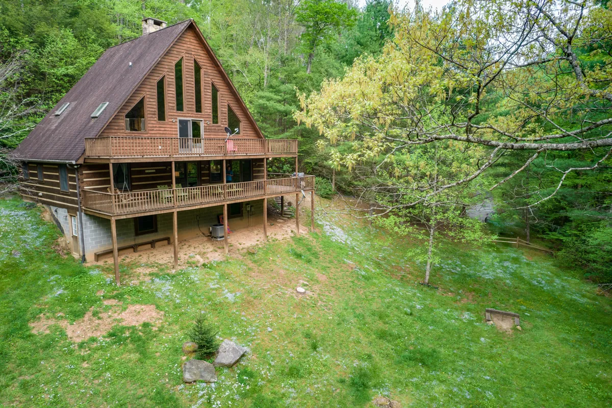 An inviting exterior shot of the A-frame cabin with a large porch surrounded by greenery and a flag on the porch. The spacious porch and open lawn suggest ample room for pets and people to enjoy the outdoors. The setting is both secluded and welcoming, ideal for relaxing after adventures in Ashe County.