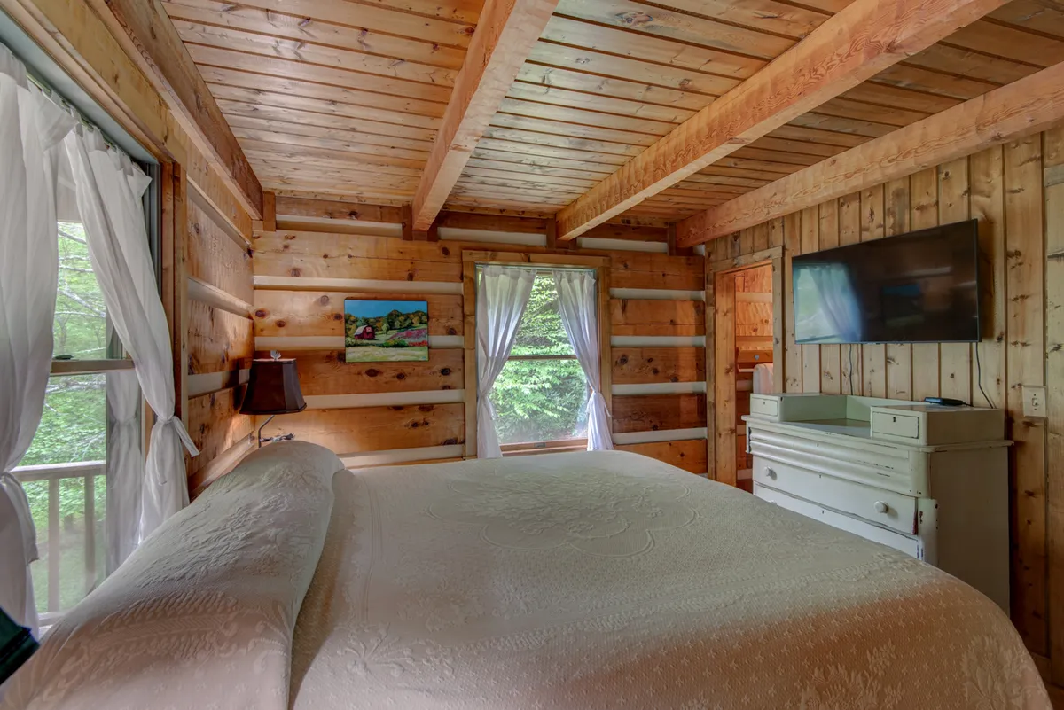 A calm and serene master bedroom featuring a comfortable bed, a large window that lets in natural light, a TV, and a dresser with artwork on the wall. The room exudes privacy and relaxation, ideal for unwinding after a day at the cabin.