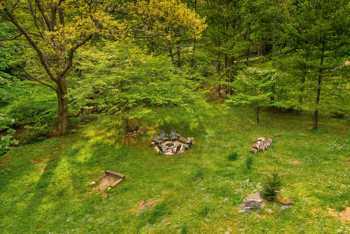 An inviting fire pit with outdoor seating sits on green grass, surrounded by tall trees. The setup is perfect for evening gatherings, s'mores, storytelling, and stargazing under the open sky.