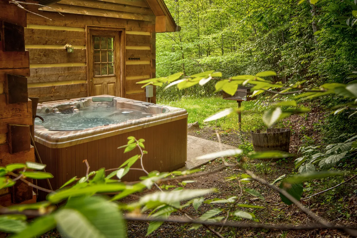 This image features an inviting hot tub nestled next to the log cabin, surrounded by lush greenery. The scene conveys a tranquil outdoor atmosphere perfect for relaxation and socializing under the open sky.