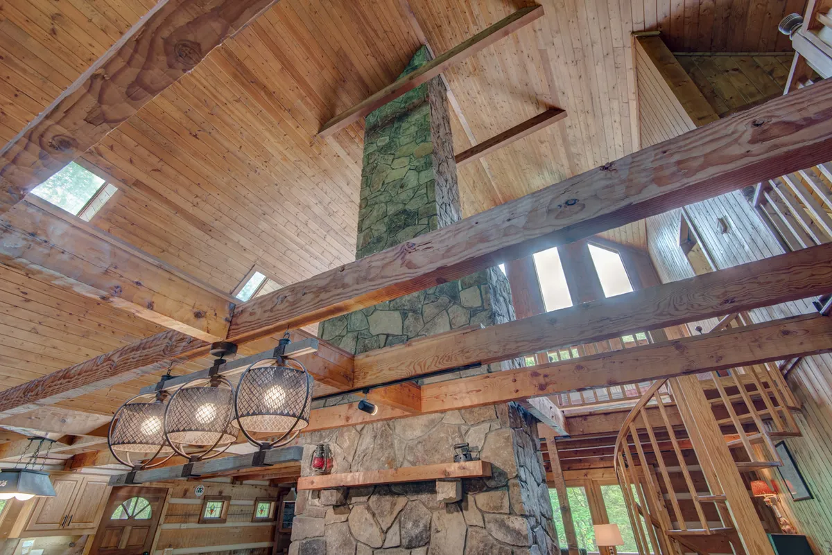 A cozy, open living space featuring a stone chimney, exposed wooden beams, large hanging light fixtures, and the spiral staircase. The openness underlines the lack of privacy and togetherness, echoing the camp-style sleepover atmosphere.